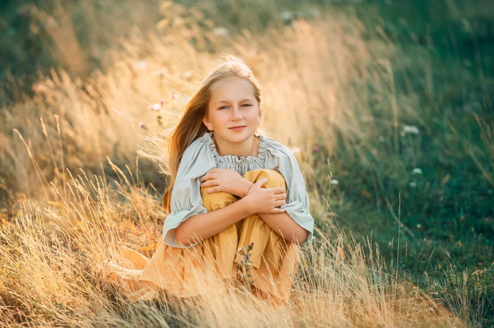 Mädchen kauert lächelnd im goldenen Abendlicht auf Wiese, Haare im Wind, warmes Fine-Art-Outdoor-Porträt